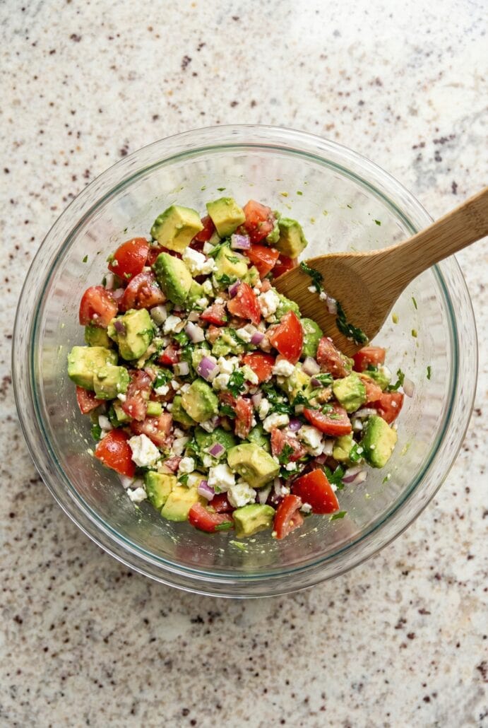 Mixing avocado feta salsa with tomatoes, red onion, herbs, and feta cheese in a glass bowl for the avocado feta salsa recipe.