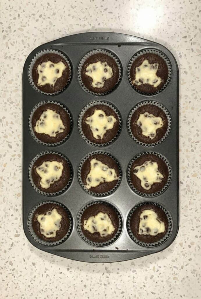 A tray of freshly baked Black Bottom Cupcakes, each with a cream cheese and chocolate chip center, showing the signature appearance of the chocolate cake with creamy filling in muffin tins.