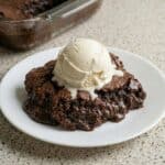A warm, fudgy chocolate brownie pudding served on a white plate and topped with a large scoop of melting vanilla ice cream.