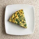 A slice of crustless spinach quiche on a white plate, showcasing a fluffy, golden egg filling with spinach and cheese.
