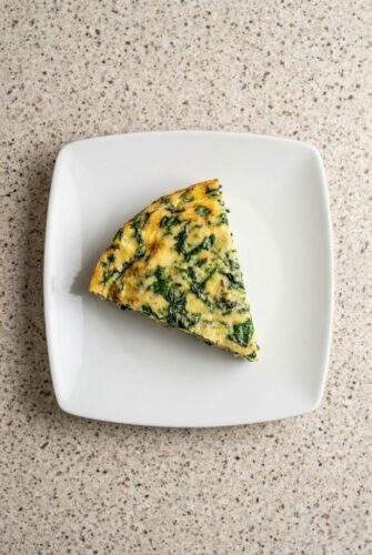 A slice of crustless spinach quiche on a white plate, showcasing a fluffy, golden egg filling with spinach and cheese.