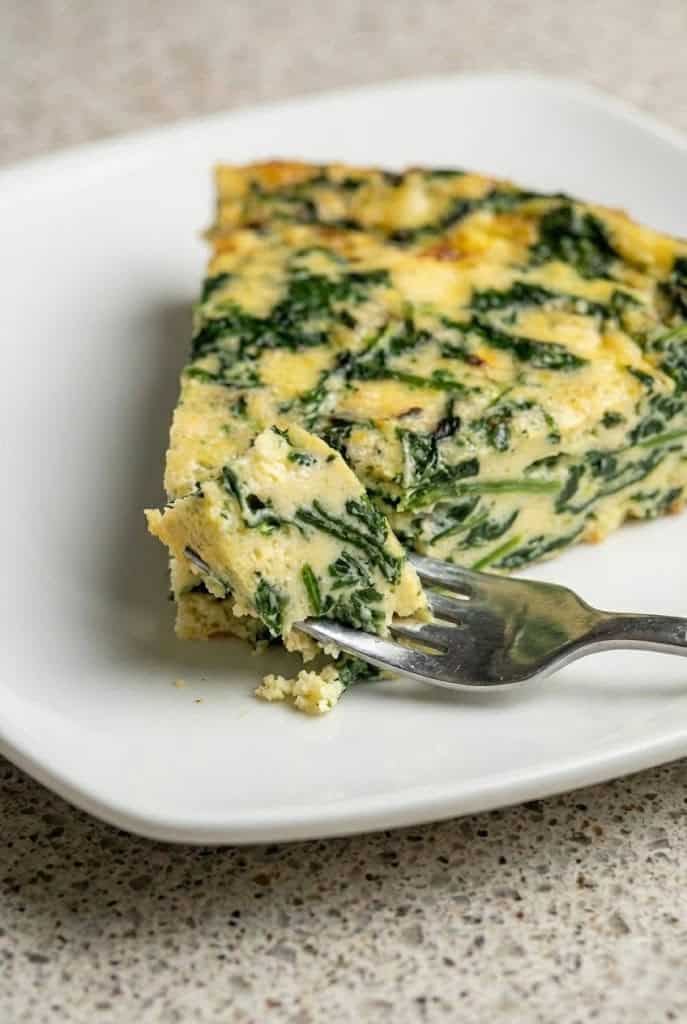 A slice of crustless spinach quiche with a fork taking a bite, highlighting the fluffy texture and vibrant green spinach filling, perfect for a healthy meal.
