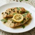 Lemon chicken piccata with capers and parsley on a white plate, topped with lemon slices.