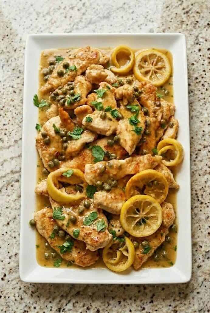 Platter of lemon chicken piccata with capers, parsley, and lemon slices.