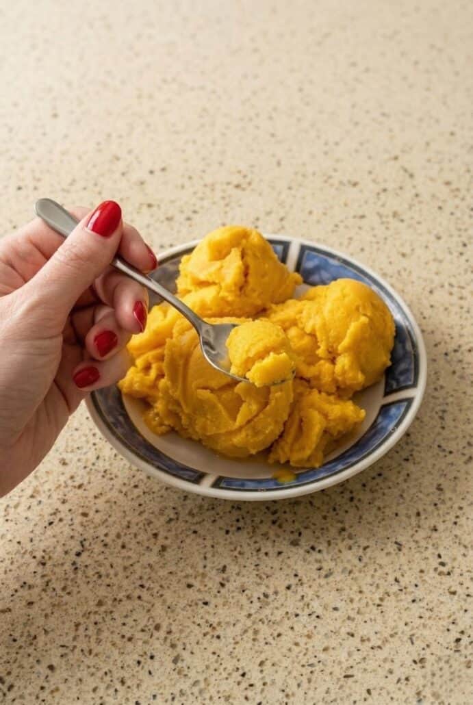 Hand holding a spoon scooping mango sorbet from a bowl, showing the creamy texture of this easy homemade mango sorbet recipe.