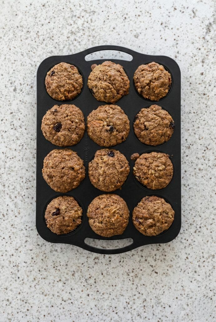 Freshly baked morning glory muffins with carrots, raisins, and nuts in a muffin pan on the counter.