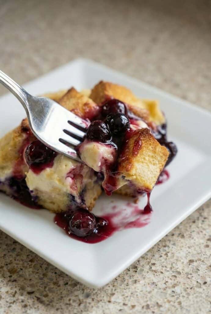 The image shows a fork taking a bite of the Overnight Blueberry French Toast Casserole, with gooey custard and fresh blueberries visible.