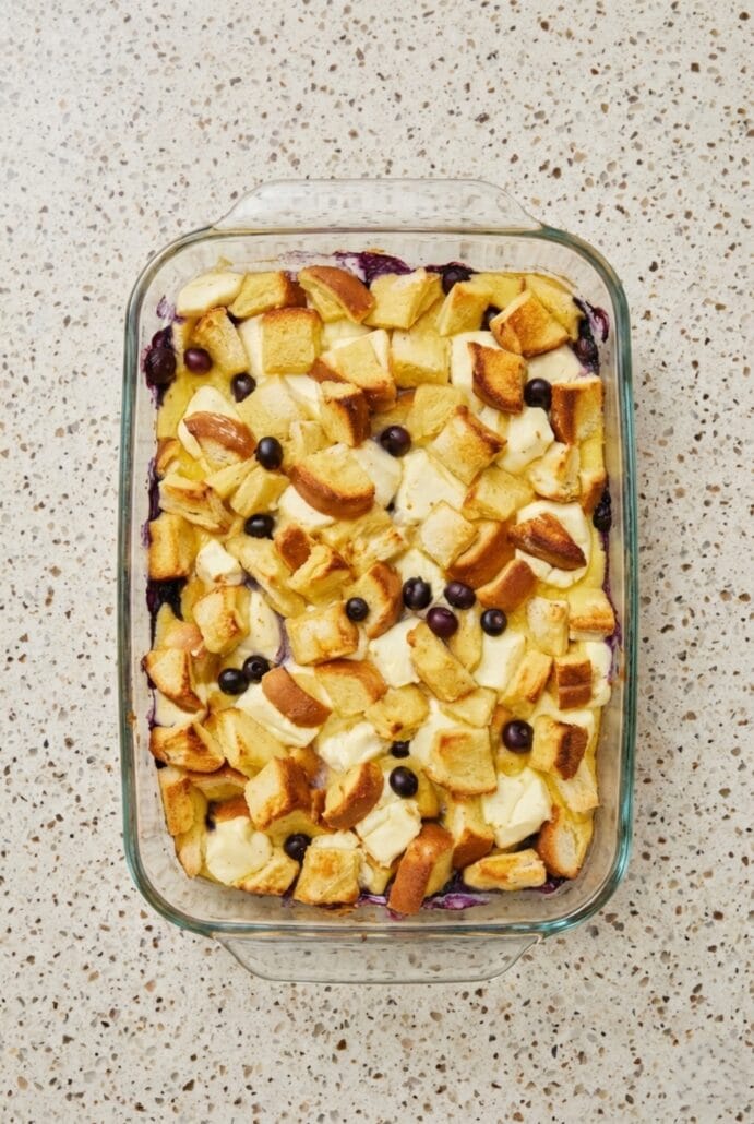 The image shows a pre-baked Overnight Blueberry French Toast Casserole in a glass baking dish, with cubed bread, cream cheese, and fresh blueberries arranged before being soaked in custard.