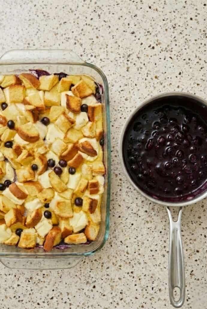 The image displays the Overnight Blueberry French Toast Casserole before baking, alongside a saucepan filled with homemade blueberry sauce.