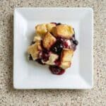 The image shows a serving of Overnight Blueberry French Toast Casserole on a white plate, drizzled with rich blueberry sauce.