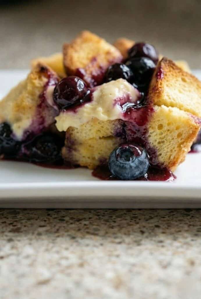 The image shows a close-up of a delicious serving of Overnight Blueberry French Toast Casserole with fresh blueberries and a rich, gooey texture from the baked custard and syrup.