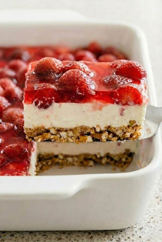 Close-up of a slice of strawberry pretzel salad being lifted from the dish, showing the crunchy pretzel crust, creamy cheesecake layer, and fresh strawberry Jell-O topping.