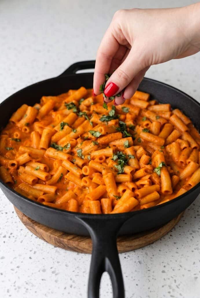 Hand sprinkling fresh basil over creamy red pepper pasta in a skillet.