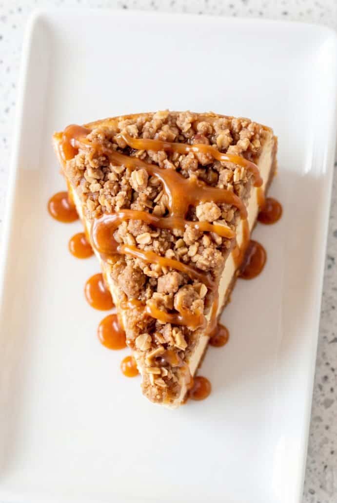 A slice of apple crisp cheesecake topped with a crunchy oatmeal crumble and drizzled with caramel sauce, highlighting the smooth, creamy layers and sweet finish.