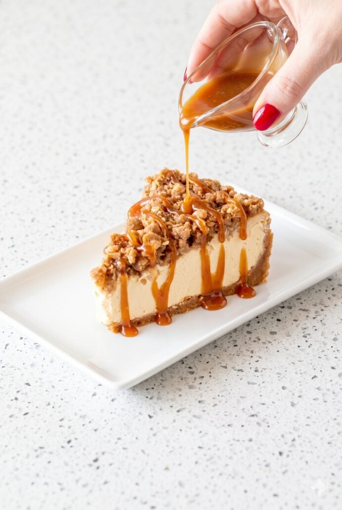 A slice of apple crisp cheesecake drizzled with caramel sauce, showcasing the crunchy topping and creamy filling.