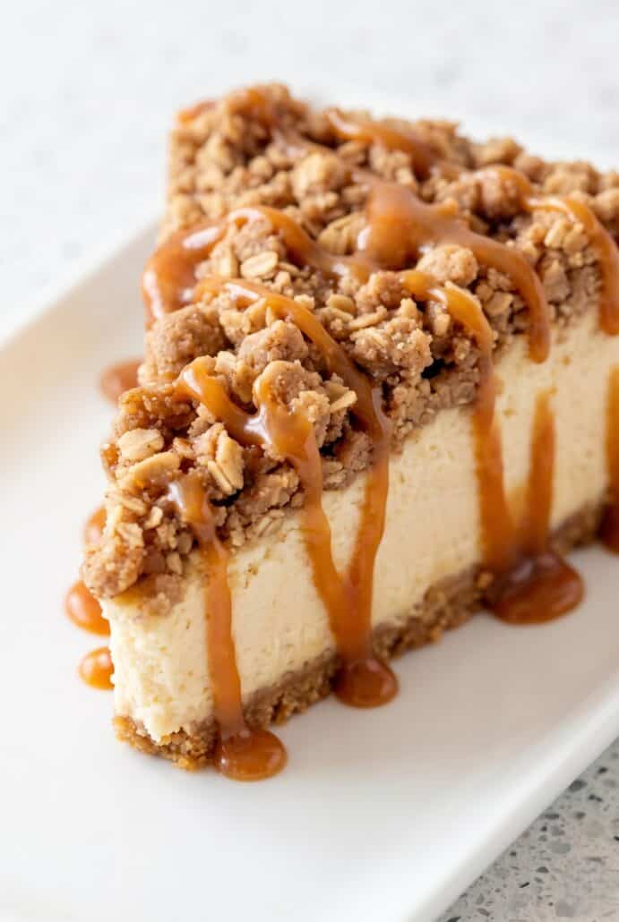 A slice of apple crisp cheesecake topped with a crunchy oatmeal crumble and drizzled with caramel sauce.