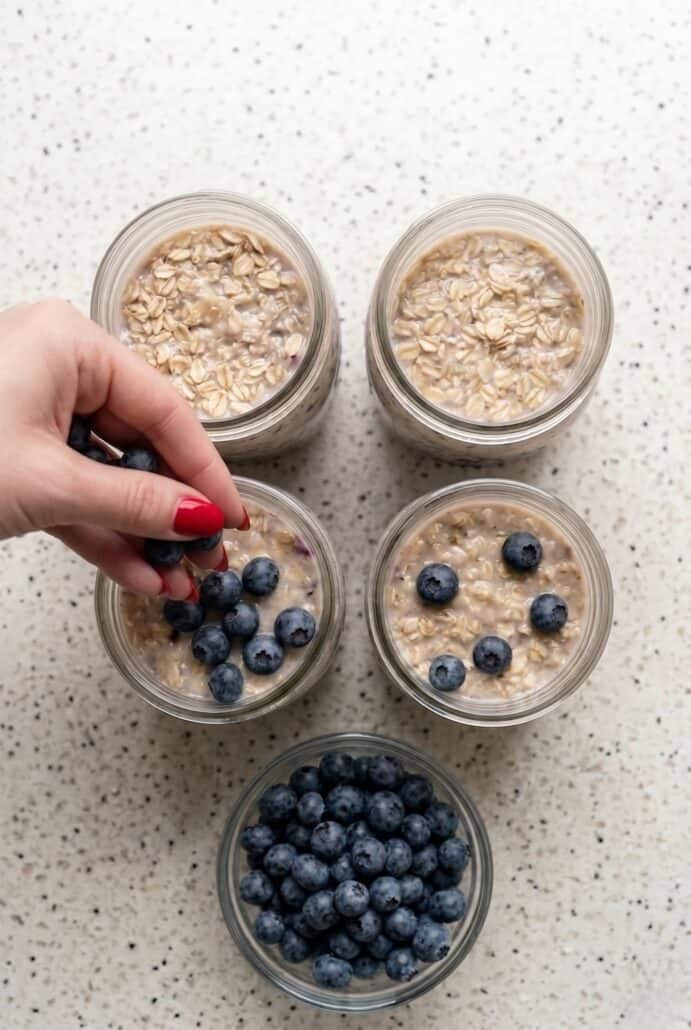 Hand adding fresh blueberries to jars of blueberry overnight oats, an easy healthy meal prep breakfast ready to enjoy.