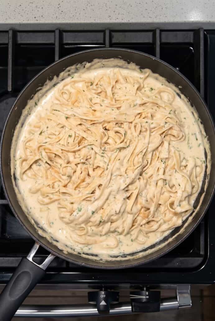 Fettuccine Alfredo pasta tossed in creamy cheese sauce cooking in a pan, shown during the sauce and pasta mixing step.