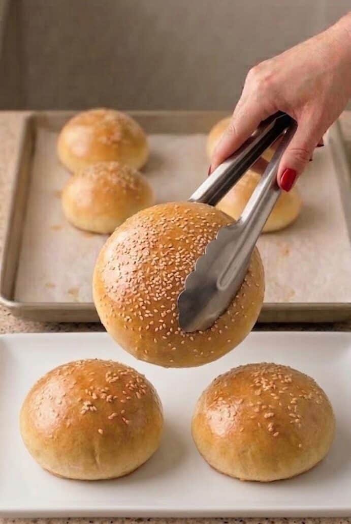 Freshly baked sesame hamburger buns being lifted from baking sheet.