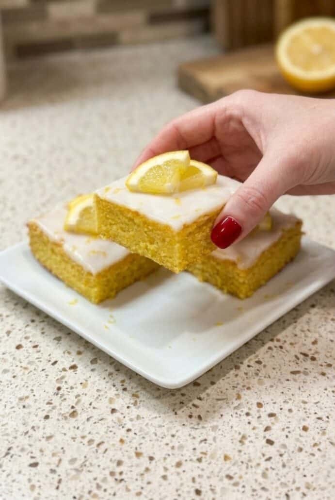 Hand holding a lemon brownie square with glaze and lemon slice topping, showing the soft texture of this easy homemade citrus dessert.