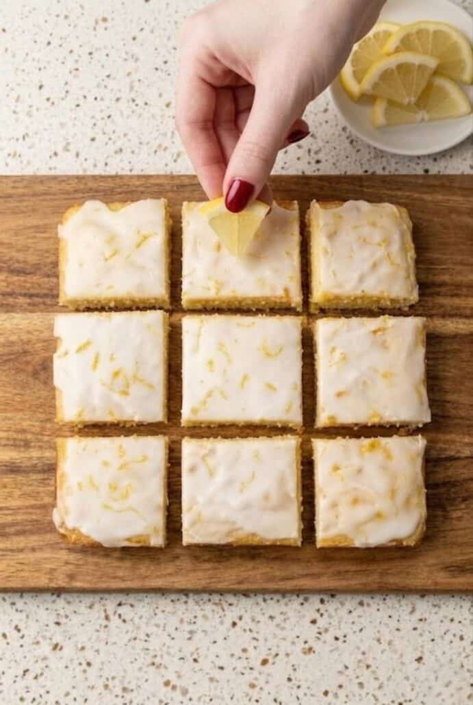 Lemon brownies cut into squares on a wooden board with glossy glaze and zest.