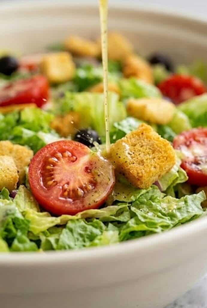 Close-up of Olive Garden salad with croutons and tomatoes being drizzled with dressing, highlighting crisp texture and fresh ingredients.