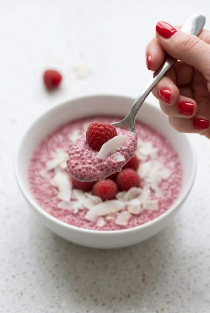 Spoon scooping raspberry chia pudding with fresh raspberries and coconut flakes, showing creamy texture.