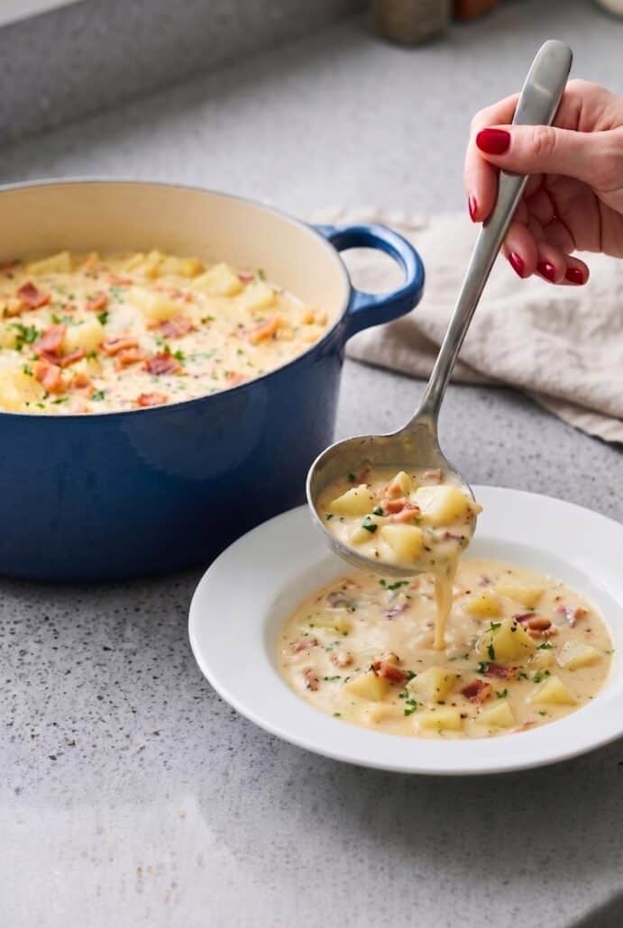 Ladle serving creamy homemade clam chowder with potatoes, bacon, and chives into a bowl.