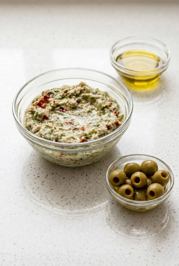 Creamy feta and green olive dip in a glass bowl with olive oil and olives on the side, perfect Mediterranean appetizer.