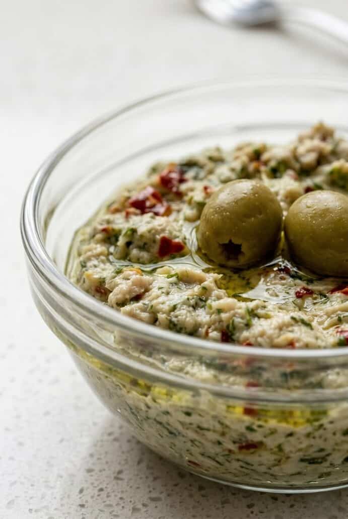 Close-up of feta and green olive spread in a glass bowl topped with olives, highlighting creamy texture.