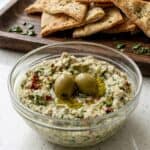 Creamy feta and green olive spread in a bowl topped with whole olives, served with pita chips.