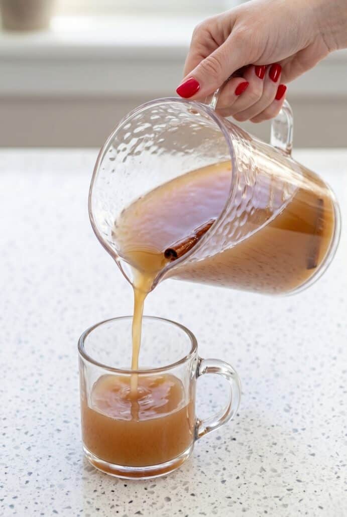Homemade apple cider being poured from a pitcher into a glass mug for serving.