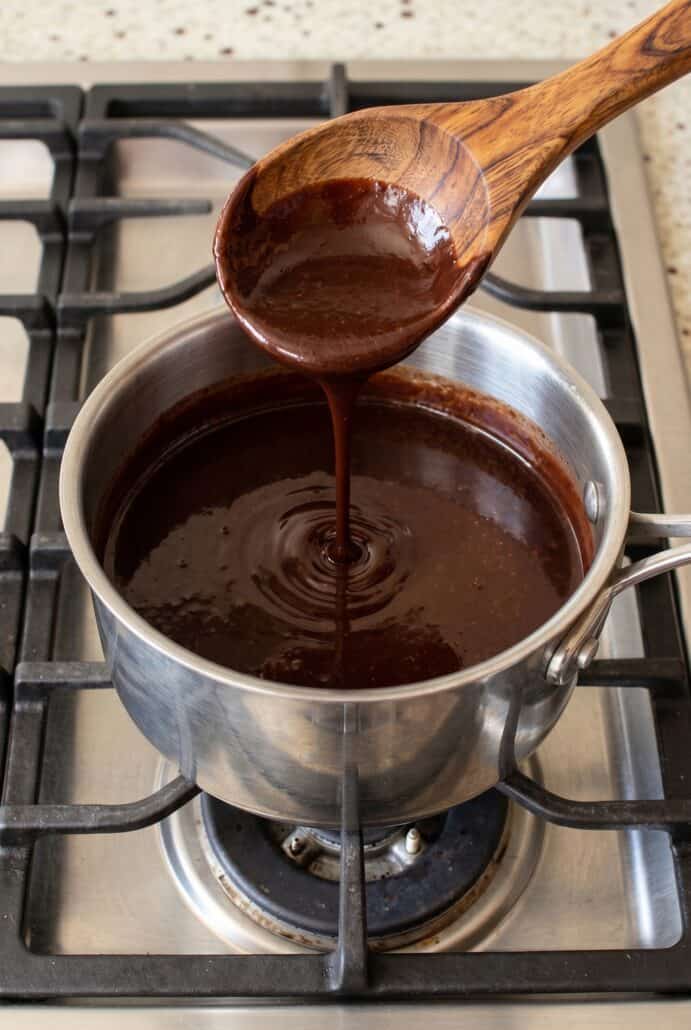 Chocolate syrup being poured into a saucepan while cooking, showing the process of making homemade chocolate syrup on the stovetop.