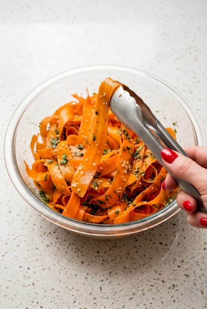Mixing raw carrot salad with thin ribbons, herbs, and sesame seeds in a bowl.
