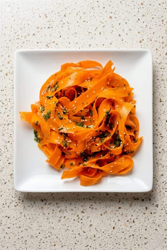 Raw carrot salad served on a white plate with carrot ribbons, herbs, and sesame seeds.