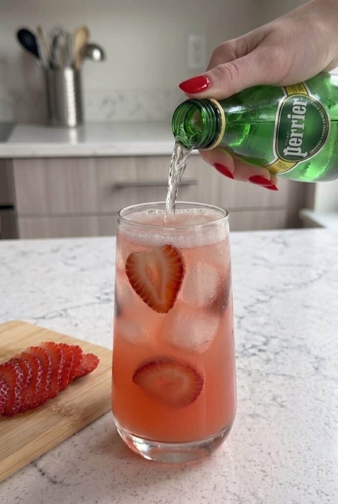 Sparkling water being poured into a glass of strawberry syrup with sliced strawberries and ice.
