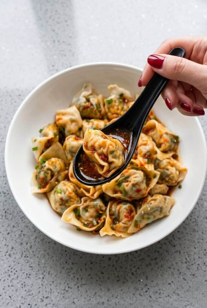 Spoon lifting vegan tofu and chive wontons with chili oil from a bowl of homemade spicy wonton dish.