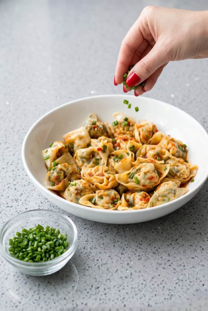 Hand sprinkling chopped chives over vegan tofu wontons with chili oil for a flavorful homemade Asian dish.