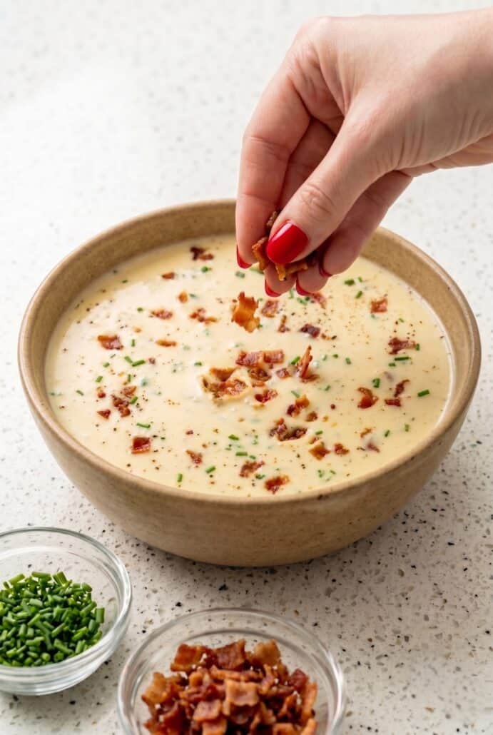 Hand sprinkling crispy bacon on creamy baked potato soup with chives.
