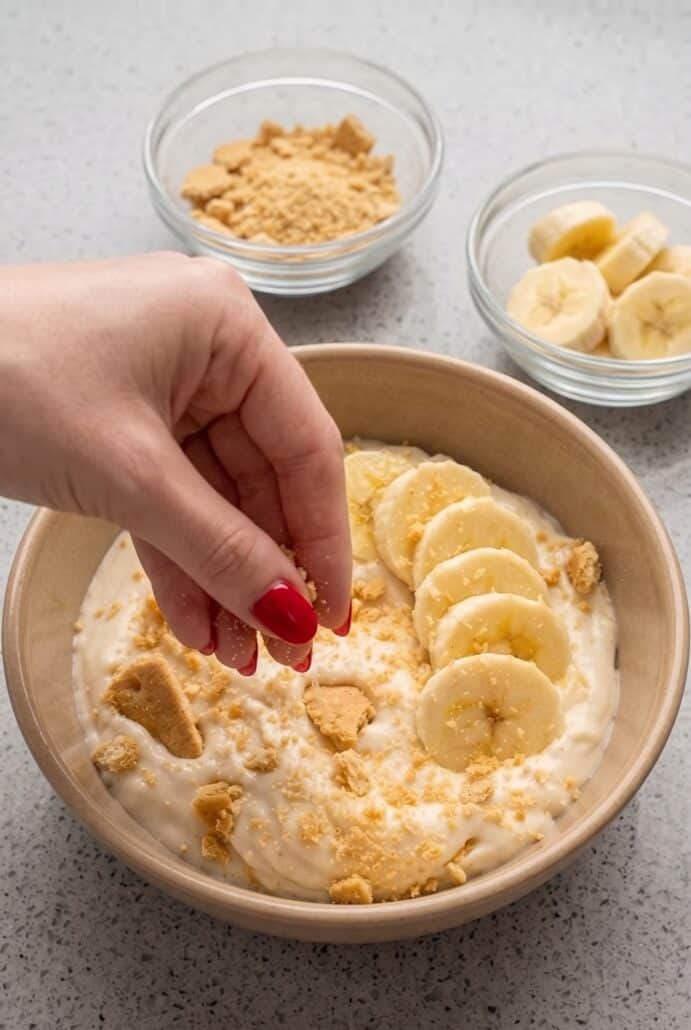 Adding graham cracker crumbs on top of creamy Banana Pudding Yogurt with fresh banana slices.