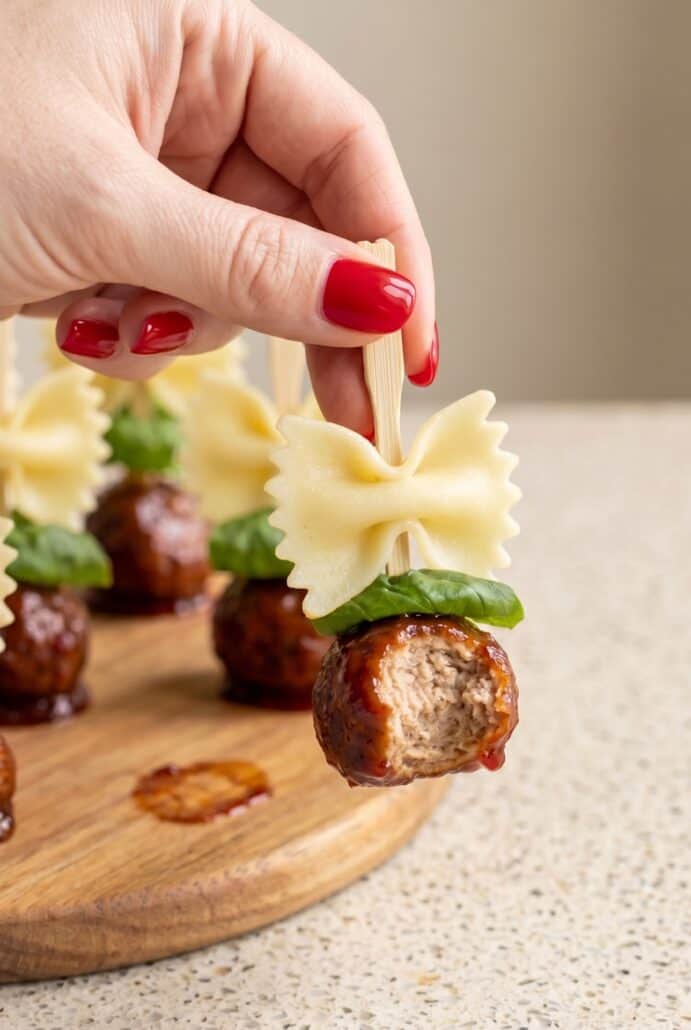 Hand holding a bowtie pasta and cocktail meatball skewer with basil for an easy party appetizer.