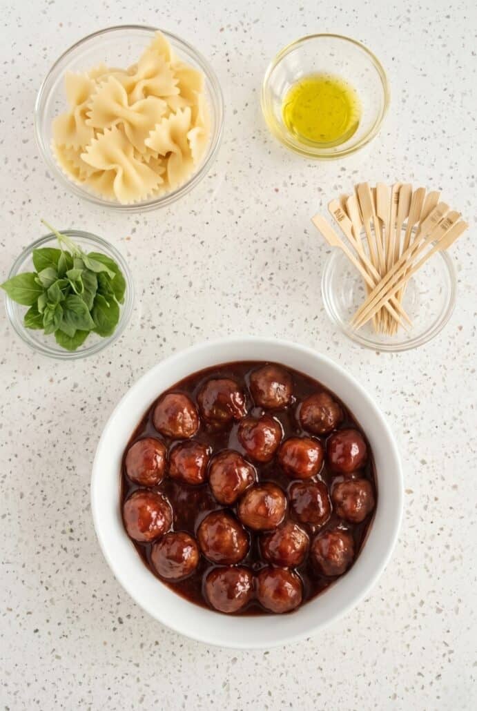 Ingredients for bowtie pasta and cocktail meatball skewers with saucy meatballs, basil, and toothpicks.