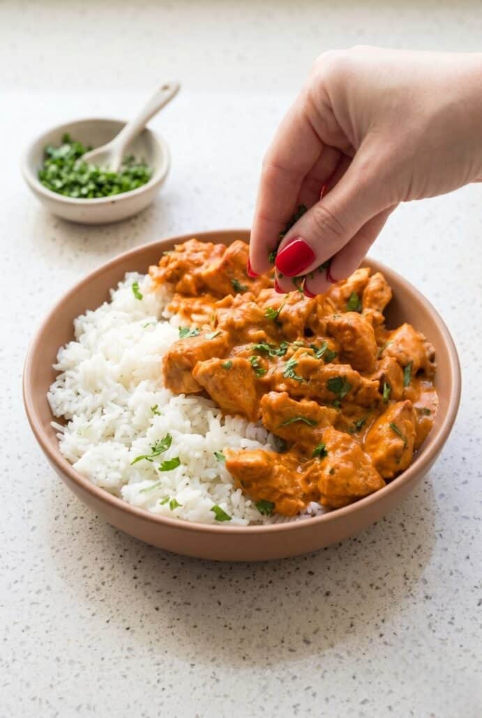 Hand garnishing coconut chicken rice bowl with fresh cilantro over white rice.