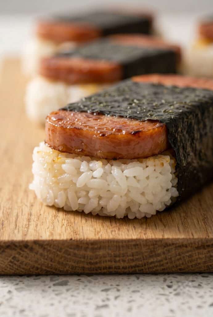 Close-up Hawaiian Spam musubi with caramelized Spam, sticky rice, and nori wrap on a wooden board.