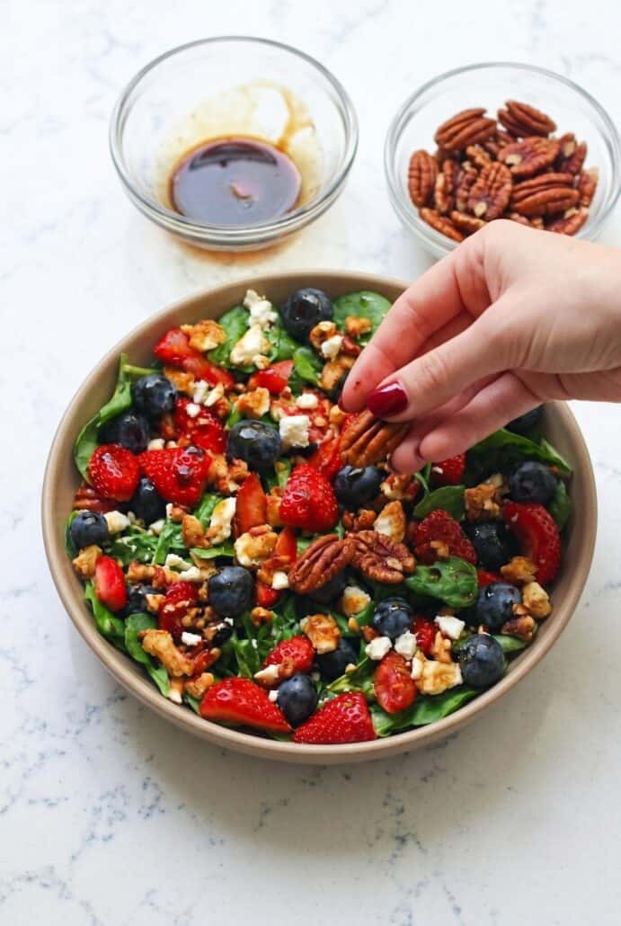 Hand sprinkling pecans over strawberry spinach salad with blueberries, feta, and balsamic glaze.