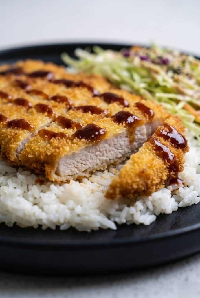 Close-up of sliced Chicken Katsu with crispy panko coating, tonkatsu sauce, rice, and cabbage slaw.