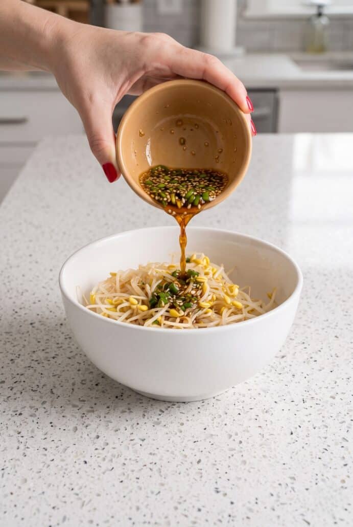 Sesame soy dressing being poured over Korean bean sprout salad in a white bowl.