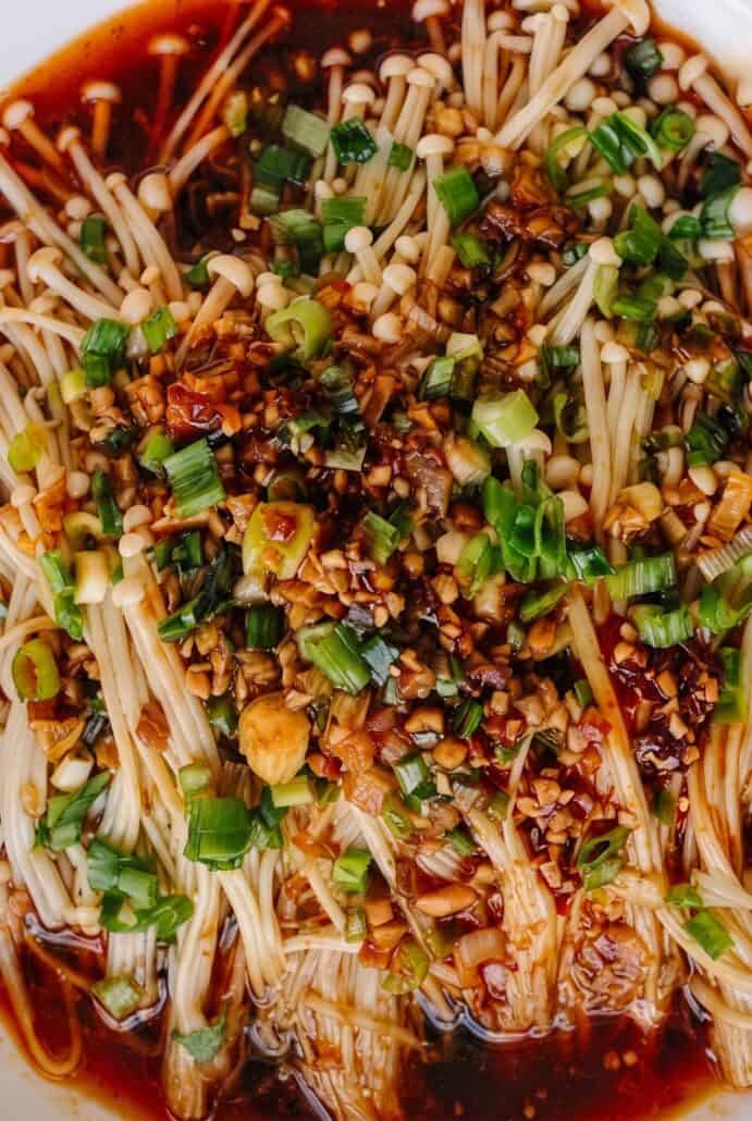 Close-up of enoki mushrooms coated in savory garlic, soy, and scallion sauce with chopped garlic and scallions on top.