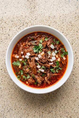 Crockpot Birria Recipe (Authentic Mexican Flavor) Mexican birria crockpot stew topped with chopped onion and cilantro in a rich red consommé.