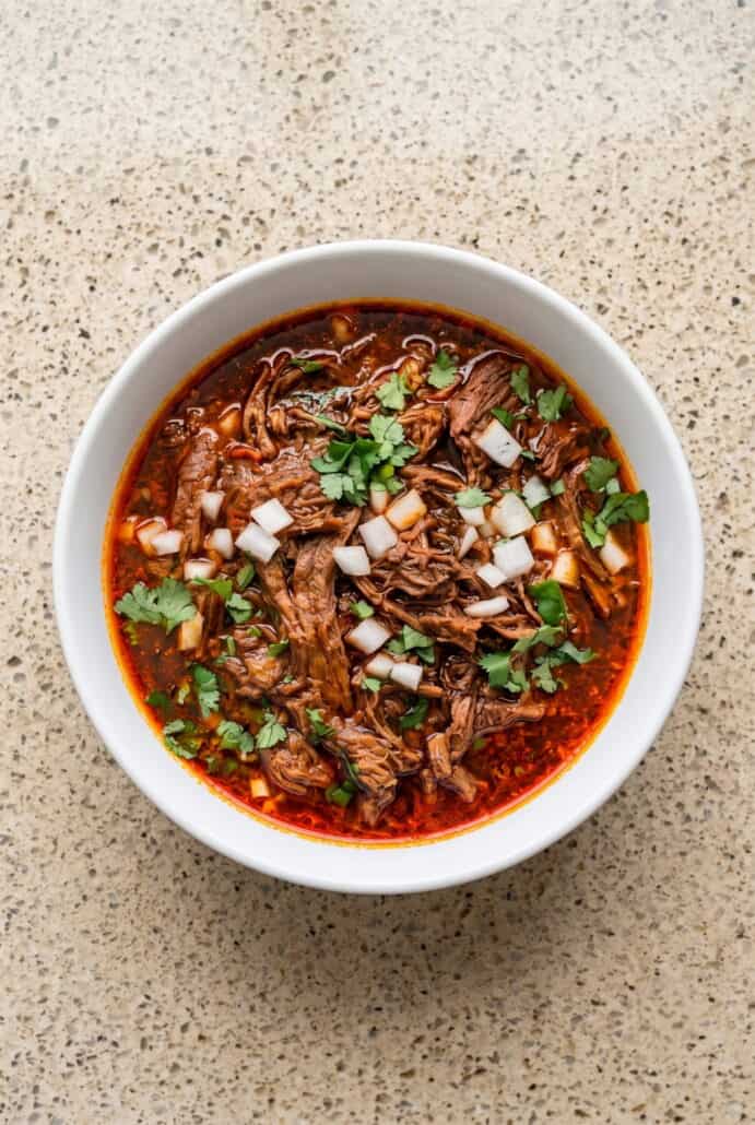 Mexican birria crockpot stew topped with chopped onion and cilantro in a rich red consommé.